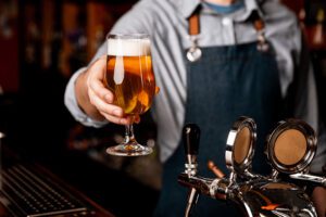 photo of a Barman who gives glass of light beer in elegant glass in dark wooden tavern interior